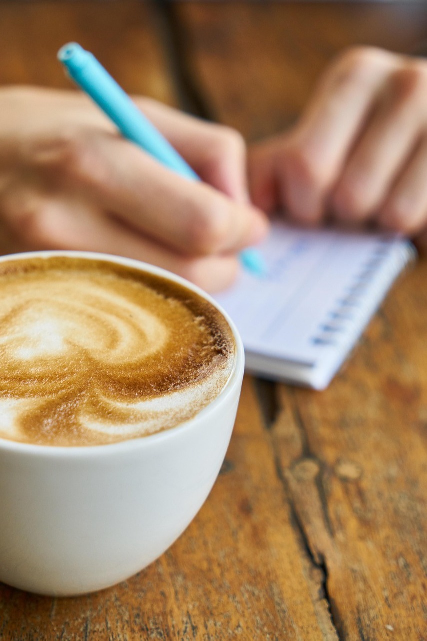 Latte art coffee on wooden table with person writing notes – planning custom orders at Wildcrafts product manufacturer.